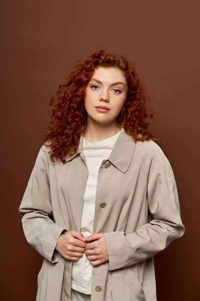 Hermosa mujer con el pelo rizado rojo posando en abrigo de otoño sobre fondo marrón, temporada de otoño - foto de stock