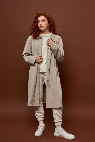 Young woman with red curly hair posing in autumn coat and joggers on brown background, fall season — Stock Photo
