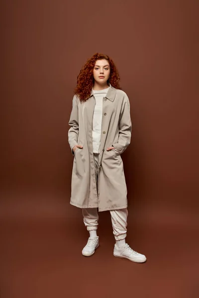 Curly woman with red hair posing in autumn coat and joggers on brown background, hands in pockets — Stock Photo
