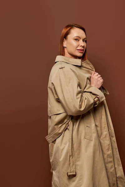 Rossa donna in trench beige guardando la fotocamera sullo sfondo marrone, moda autunno stagione — Foto stock