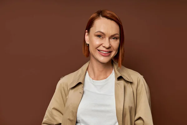 Heureuse rousse femme en trench coat beige regardant la caméra sur fond brun, mode automne — Photo de stock