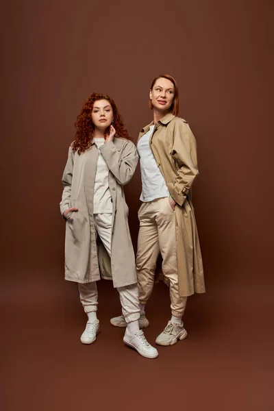 Two generations, redhead women in trendy autumn clothes posing on brown background, trench coats — Stock Photo