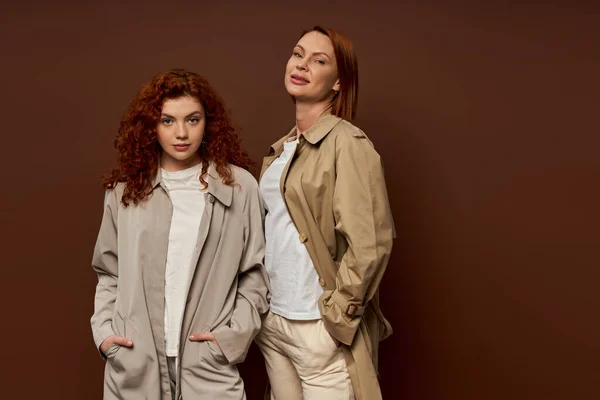 Two generations, redhead women in trendy autumn attire posing on brown background, trench coats — Stock Photo
