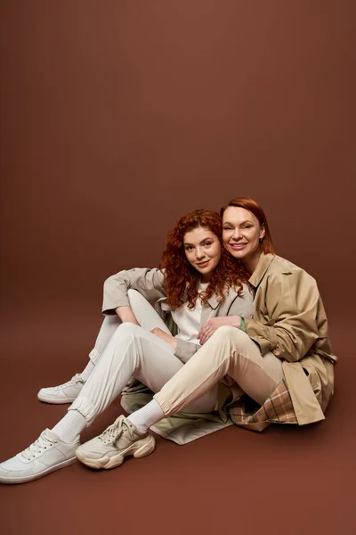 Deux générations, femmes positives aux cheveux roux posant en trench d'automne sur fond brun — Photo de stock