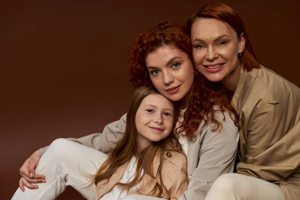 Tre generazioni di donne, felice famiglia rossa guardando la fotocamera e sorridendo su sfondo marrone — Foto stock