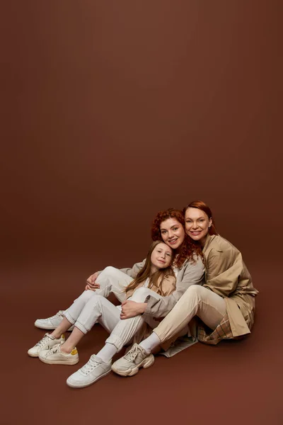 Trois générations, famille rousse positive regardant la caméra sur fond brun, les femmes et les enfants — Photo de stock
