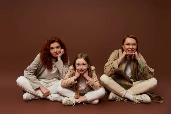 Tres generaciones de mujeres mirando a la cámara y sentado en abrigos en el fondo marrón, linda chica - foto de stock