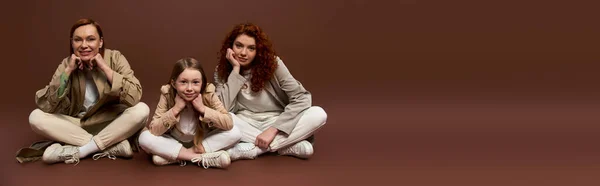 Three generations of women looking at camera and sitting in coats on brown backdrop, banner — Stock Photo