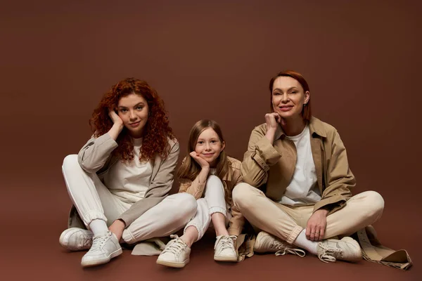Felice famiglia rossa di tre generazioni femminili seduti su sfondo marrone, cappotti autunnali — Foto stock