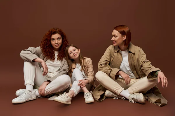 Cheerful redhead family of three female generations sitting on brown background, autumn coats — Stock Photo