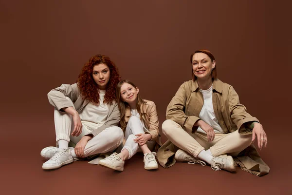 Alegre familia pelirroja de tres generaciones femeninas sentadas sobre fondo marrón, moda otoñal - foto de stock