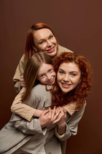 Familia pelirroja feliz de tres generaciones femeninas en abrigos abrazándose unos a otros sobre fondo marrón - foto de stock