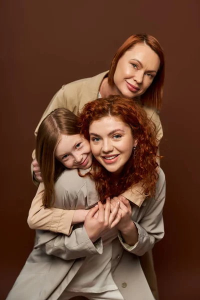 Alegre familia pelirroja de tres generaciones femeninas en abrigos abrazándose unos a otros sobre fondo marrón - foto de stock