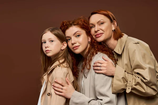 Familia de tres generaciones femeninas con el pelo rojo posando en abrigos beige, abrazándose sobre fondo marrón - foto de stock