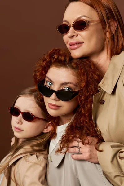 Family of three female generations with red hair posing in sunglasses and coats on brown backdrop — Stock Photo