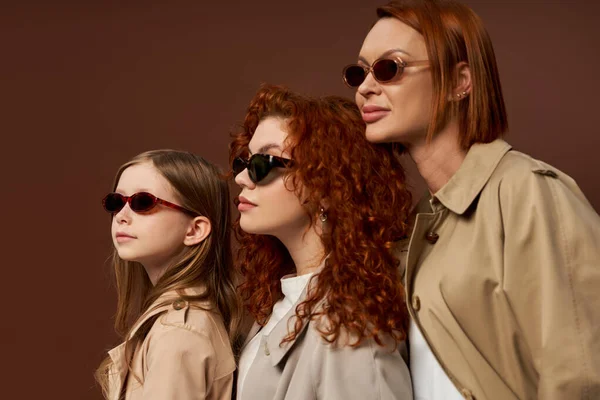 Three female generations with red hair posing in sunglasses and coats on brown backdrop, family bond — Stock Photo