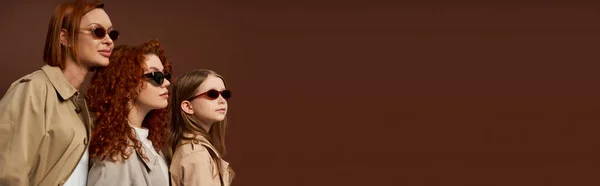 Tre generazioni femminili con capelli rossi in posa in occhiali da sole e cappotti su sfondo marrone, striscione — Foto stock