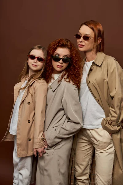 Three female generations with red hair posing in sunglasses and coats on brown backdrop, family bond — Stock Photo