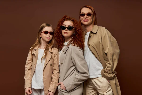 Три жіночі покоління, жінки і дитина з червоним волоссям позує в сонцезахисних окулярах і пальто на коричневому — стокове фото