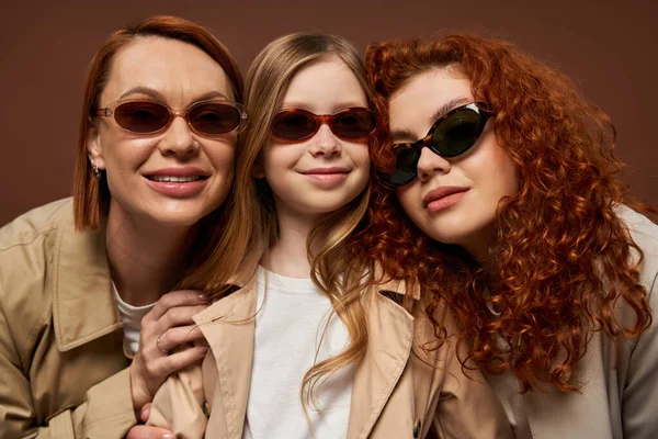 Concepto de cambio de generación familiar, mujeres pelirroja feliz y chica en gafas de sol sobre fondo marrón - foto de stock
