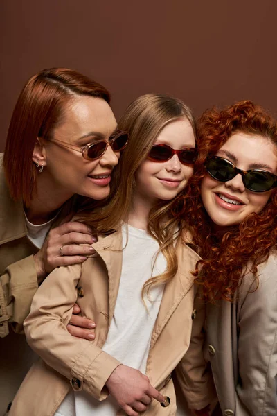 Family generation change concept, cheerful redhead women and girl in sunglasses on brown backdrop — Stock Photo