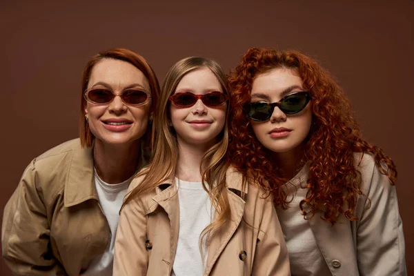 Портрет счастливых трех поколений женщин, смотрящих на камеру в солнечных очках на коричневом фоне — стоковое фото