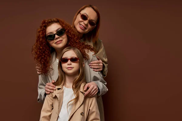 Семейный портрет трех поколений женщин в солнечных очках и пальто, позирующих на коричневом фоне — стоковое фото