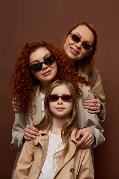 Сімейний портрет трьох жіночих поколінь в сонцезахисних окулярах і пальто, посміхаючись на коричневому фоні — стокове фото