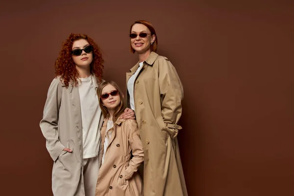 Portrait de famille de générations féminines en lunettes de soleil et manteaux sur fond brun, les mains dans les poches — Photo de stock