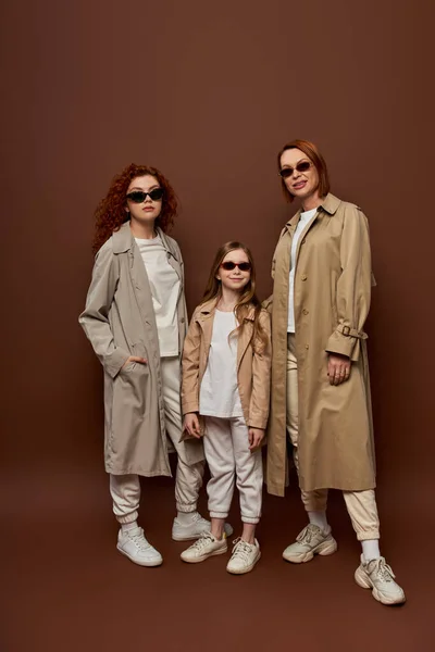 Photo de famille de générations féminines en lunettes de soleil et manteaux posant sur fond brun, pleine longueur — Photo de stock