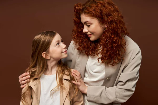 Два покоління, молода кучерява мати з рудим волоссям дивиться на дочку на коричневому фоні, сім'я — стокове фото
