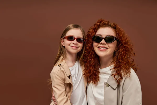 Два покоління, щаслива кучерява мати і дочка в сонцезахисних окулярах на коричневому тлі, портрет — стокове фото