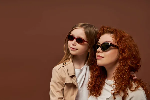 Deux générations, heureuse rousse mère et fille en lunettes de soleil regardant loin sur fond brun — Photo de stock