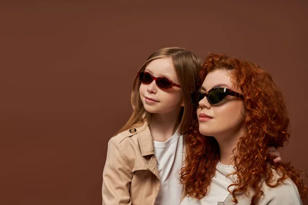 Dos generaciones, feliz pelirroja madre e hija en gafas de sol mirando hacia otro lado en el fondo marrón - foto de stock