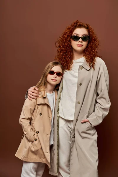 Herbst-Stil, rothaarige Mutter und Kind posieren in Sonnenbrille und beigem Trenchcoat, brauner Hintergrund — Stockfoto