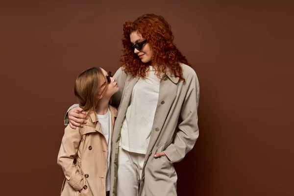 Autumn fashion, redhead mother and kid posing in sunglasses and beige trench coats, brown backdrop — Stock Photo