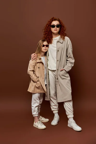 Generaciones, madre pelirroja y el niño posando en gafas de sol y abrigos de otoño beige, fondo marrón - foto de stock