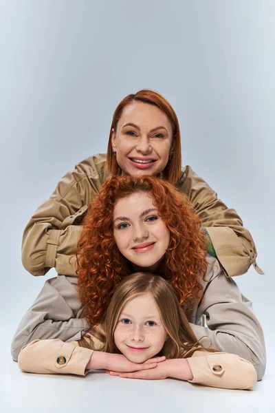 Happy three generations, redhead mother and child in beige trench coats posing on grey background — Stock Photo