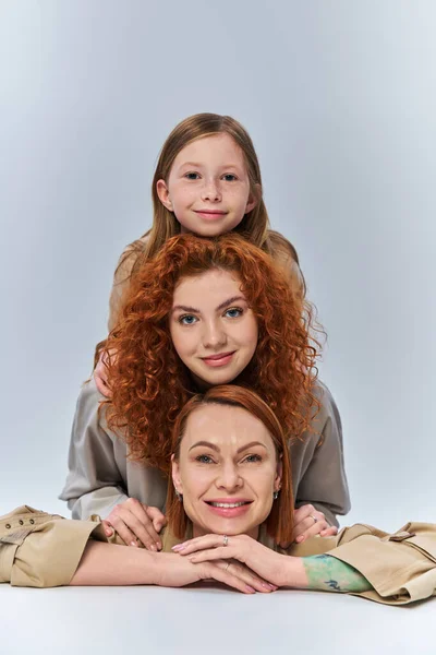 Trois générations, heureuse famille de rousses en manteaux beiges souriant sur fond gris, lien féminin — Photo de stock
