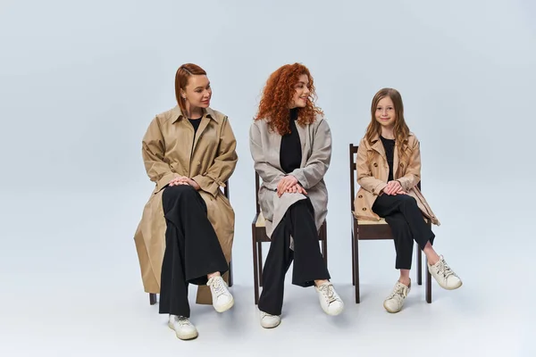 Tre generazioni, donne rosse in cappotti sedute su sedie e guardando la ragazza sullo sfondo grigio — Foto stock