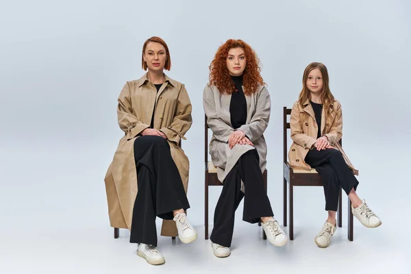 Three female generations, redhead women and child in coats sitting on chairs on grey backdrop — Stock Photo