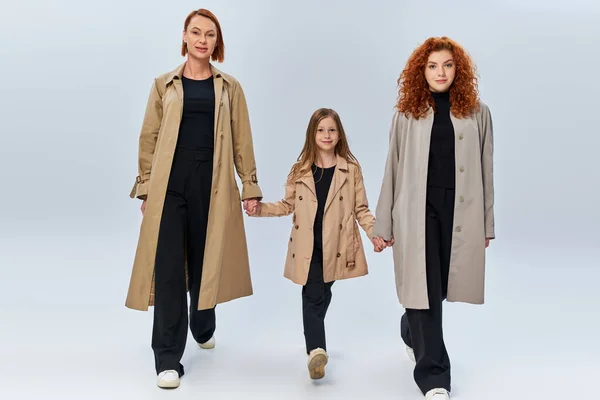 Tre generazioni femminili, donne rosse e ragazza in cappotti che si tengono per mano e camminano su sfondo grigio — Foto stock