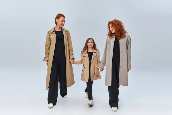 Drei Generationen, rothaarige Frauen und Mädchen in Trenchcoats, Händchen haltend und vor grauem Hintergrund wandelnd — Stockfoto