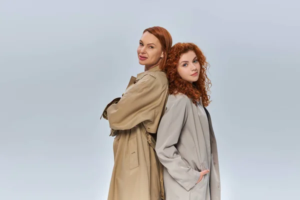Два покоління рудих жінок, що позують в автентичних траншеях пальто на сірому фоні, сезонний одяг — стокове фото