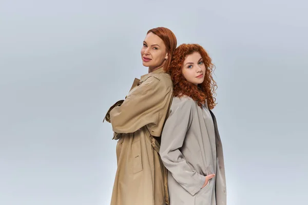 Dos generaciones de mujeres pelirrojas posando en gabardinas otoñales sobre fondo gris, atuendo estacional - foto de stock