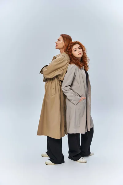 Dos generaciones de mujeres con el pelo rojo de pie en gabardinas sobre fondo gris, moda otoñal - foto de stock