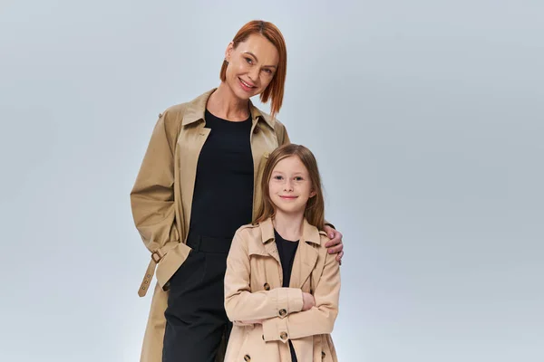 Two generations, woman with red hair and little girl standing in trench coats on grey background — Stock Photo