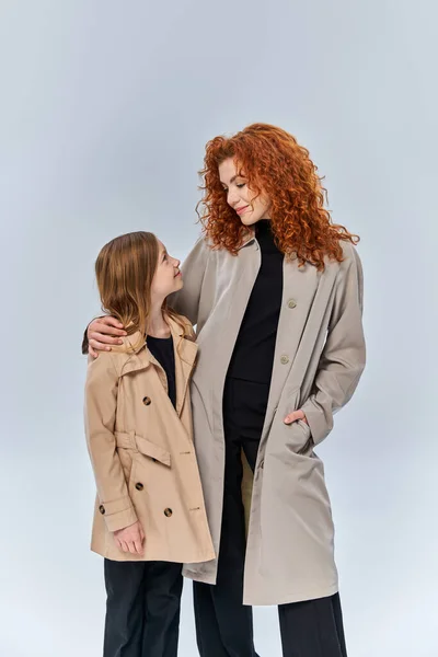 Dos generaciones femeninas, pelirroja mirando a su hija y de pie con abrigos sobre fondo gris - foto de stock