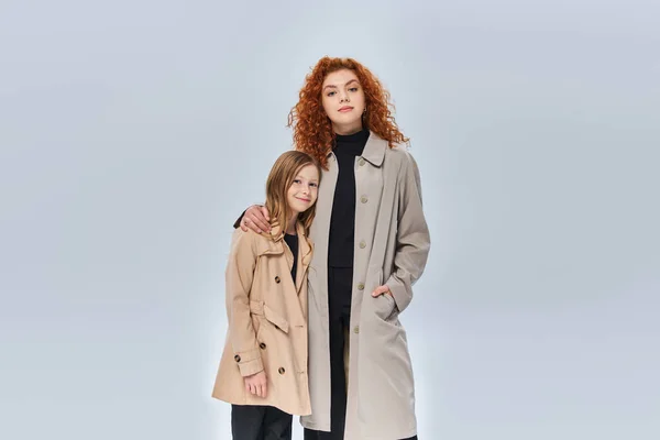 Dos generaciones femeninas, pelirroja abrazando a su hija y de pie con abrigos sobre fondo gris — Stock Photo