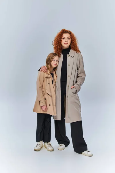 Dos generaciones femeninas, pelirroja abrazando a un niño feliz y de pie con abrigos sobre fondo gris - foto de stock